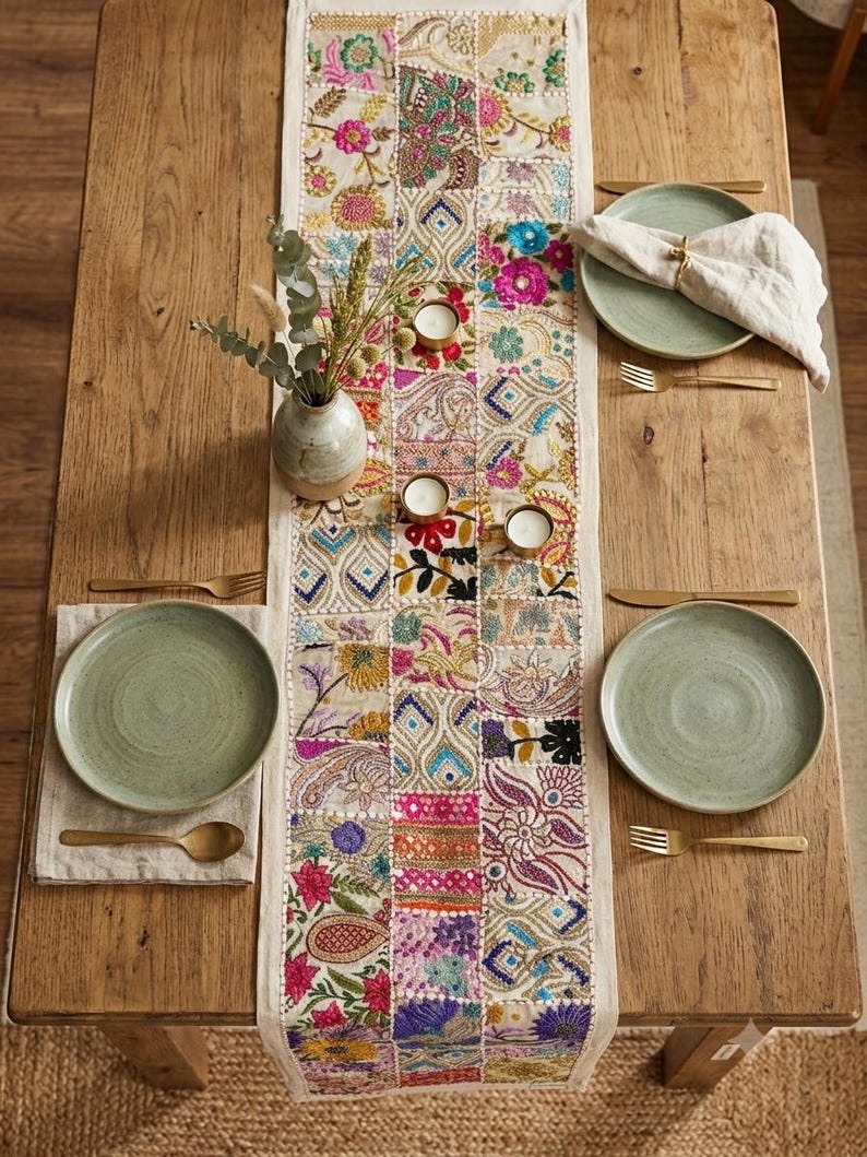 May include: A rectangular table runner featuring a patchwork design of embroidered fabric squares in vibrant colors including pink, blue, yellow, and green. The runner is centered on a wooden table with four green plates, gold cutlery, and candles.
