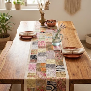 May include: A rectangular table runner featuring a patchwork design of various embroidered fabric squares in shades of pink, yellow, green, and black. The runner is centred on a wooden table with two terracotta plates and beige napkins.