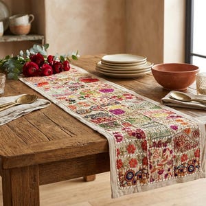 May include: A rectangular table runner featuring a patchwork design of embroidered floral and geometric patterns in vibrant colors like red, pink, yellow, and blue on a light beige fabric base, draped across a rustic wooden dining table.