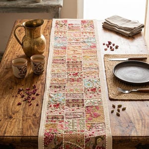 Può includere: Un runner da tavola rettangolare caratterizzato da un design patchwork di quadrati di tessuto ricamati con vari motivi floreali e geometrici, arricchito da paillettes e perline, disposto su un tavolo in legno con una brocca in ottone e tazze.