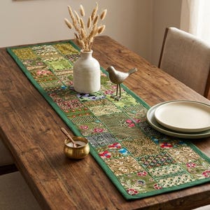 Peut inclure: Un chemin de table rectangulaire pr&eacute;sentant un motif patchwork compos&eacute; de divers carr&eacute;s de tissu brod&eacute;s dans des tons verts, dor&eacute;s et floraux. Le chemin de table poss&egrave;de une bordure verte unie et est orn&eacute; de broderies complexes et de fils m&eacute;talliques.