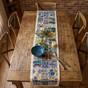 Puede incluir: Un camino de mesa rectangular con un diseño de patchwork de varios patrones florales y geométricos en azul, verde, amarillo y blanco. El camino está centrado en una mesa de comedor de madera con cuatro sillas, un cuenco azul y cubiertos.