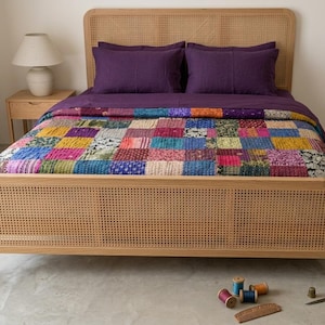 May include: A bed with a light wood frame and headboard, featuring a woven cane design. The bed is made up with purple sheets and a colorful patchwork quilt. A small side table with a lamp is visible in the background.