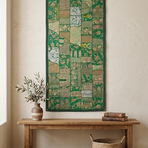 May include: A rectangular wall hanging featuring a patchwork of green and gold fabric panels with intricate floral and geometric patterns. The textile is bordered in dark green fabric and includes metallic thread accents throughout the design.