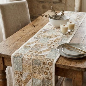 May include: A rectangular table runner featuring a patchwork design of embroidered fabric squares in shades of cream, beige, sage green, and gold. The runner has a solid cream border and is laid flat across a wooden dining table with place settings.