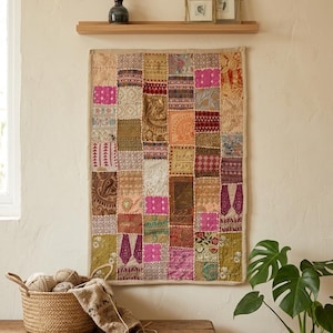 May include: A rectangular wall hanging composed of various patchwork fabric squares in shades of pink, gold, green, and brown. The textile features intricate embroidery, metallic threads, and diverse patterns, finished with a simple beige fabric border.