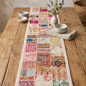 May include: A rectangular table runner featuring a patchwork design of various embroidered fabric squares in vibrant colors including pink, gold, blue, and cream. The runner has a white fabric border and is decorated with intricate floral and geometric patterns.