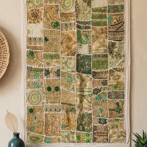 Puede incluir: Un tapiz de pared patchwork con una cuadrícula de cuadrados de tela en tonos verdes, beige y crema. Los cuadrados muestran varios patrones, incluyendo diseños florales y paisley. Los bordes están cosidos con hilo blanco, y la pieza está enmarcada con un borde crema.