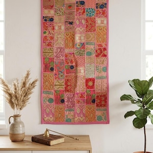 May include: A rectangular wall hanging featuring a patchwork design composed of various fabric squares in shades of pink, orange, red, and gold. The textile displays intricate embroidered patterns, metallic thread accents, and a solid pink border.