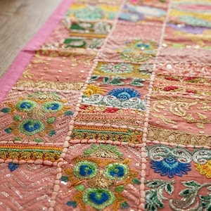 Pode incluir: Um caminho de mesa retangular com um design de patchwork em pain&eacute;is cor-de-rosa. Cada sec&ccedil;&atilde;o &eacute; decorada com bordados intrincados em fios azul, verde, amarelo e dourado, acentuados com pequenas lantejoulas refletoras e costuras met&aacute;licas.