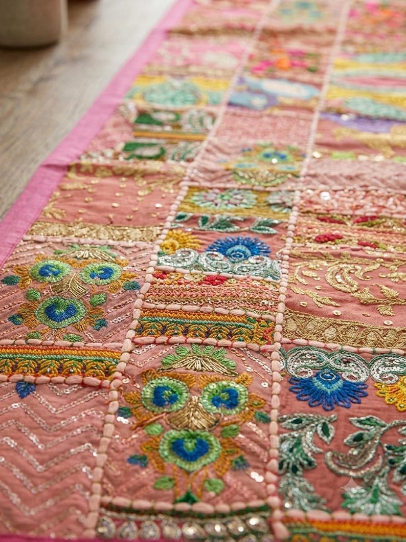 Pode incluir: Um caminho de mesa retangular com um design de patchwork em pain&eacute;is cor-de-rosa. Cada sec&ccedil;&atilde;o &eacute; decorada com bordados intrincados em fios azul, verde, amarelo e dourado, acentuados com pequenas lantejoulas refletoras e costuras met&aacute;licas.