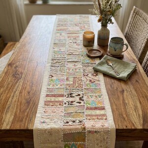 Peut inclure: Chemin de table rectangulaire en patchwork aux couleurs beige, rose et bleu, posé sur une table en bois. L'image présente également un vase de fleurs séchées, une bougie et une tasse. Le motif du chemin de table est fait de carrés de tissu.