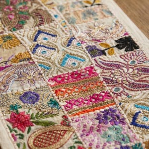 May include: A rectangular fabric runner featuring a patchwork design of embroidered floral and geometric patterns. The textile displays vibrant threads in pink, purple, blue, green, and gold, accented with small reflective circular mirrors.