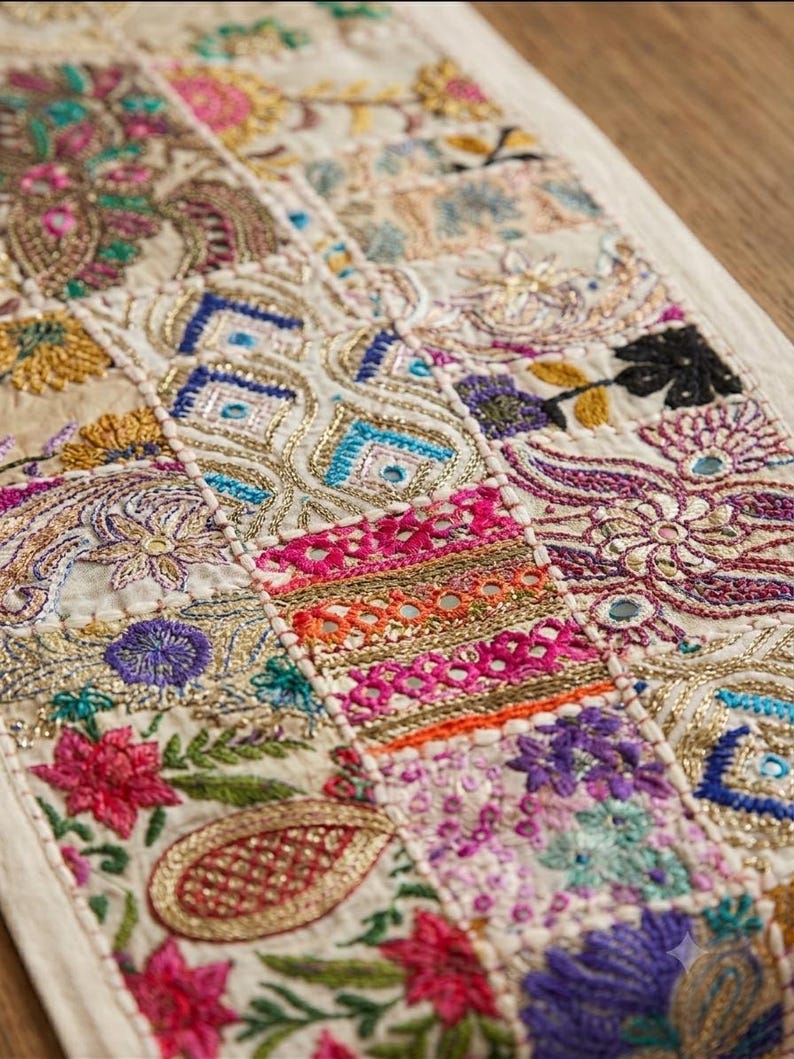May include: A rectangular fabric runner featuring a patchwork design of embroidered floral and geometric patterns. The textile displays vibrant threads in pink, purple, blue, green, and gold, accented with small reflective circular mirrors.