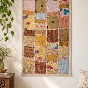 Peut inclure: Une tenture murale rectangulaire composée d'un assemblage de carrés de tissus variés dans des tons dorés, roses, bleus et marron. Le textile présente des broderies complexes, des motifs floraux et des pompons entre les sections.