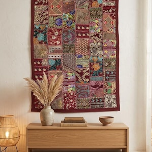 Puede incluir: Un tapiz de pared decorativo de patchwork con un borde burdeos. El diseño presenta una variedad de cuadrados estampados en tonos de rojo, dorado, azul y verde. El tapiz se exhibe sobre un mueble de madera con objetos decorativos.