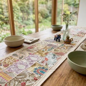 May include: A rectangular table runner featuring a patchwork design of embroidered fabric squares in various colours including gold, pink, blue, and white. The embroidery includes floral and paisley patterns with raised textures and decorative stitching.