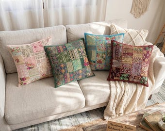 Op de afbeelding: Vier vierkante sierkussens met complexe patchworkontwerpen in tinten roze, groen, blauwgroen en donkerrood. Elk kussen toont een unieke opstelling van geborduurde stoffen vierkanten met gevarieerde patronen, texturen en decoratieve stiksels.