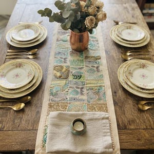 May include: A wooden dining table set for four with cream-colored plates featuring floral patterns, gold-toned cutlery, a copper vase with dried roses, and a rectangular table runner with a patchwork design in blue, green, and gold tones.
