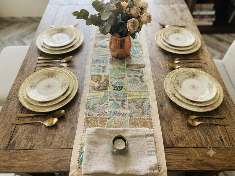 May include: A wooden dining table set for four with cream-colored plates featuring floral patterns, gold-toned cutlery, a copper vase with dried roses, and a rectangular table runner with a patchwork design in blue, green, and gold tones.