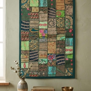 May include: A patchwork textile wall hanging with a dark green border. The squares are various shades of green, blue, and gold, with floral and geometric patterns. The textile is hung with small wooden clips.