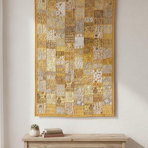 Puede incluir: Un tapiz rectangular con un diseño de patchwork en tonos amarillos, dorados y crema. La pieza, similar a un edredón, presenta una variedad de cuadrados estampados y está enmarcada con un borde amarillo. Debajo hay un banco de madera con libros y una planta.