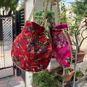 May include: A collection of embroidered drawstring bags in vibrant colors. The bags are red, pink, and green, adorned with floral patterns and gold accents. Each bag hangs from a beaded strap, showcasing a blend of textures and hues.