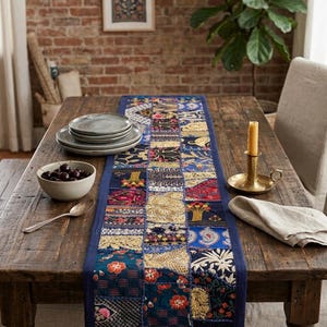May include: A rectangular table runner featuring a patchwork design of various blue, gold, and red fabric squares with intricate embroidery, metallic thread detailing, and floral patterns, centered on a wooden dining table with a blue border.