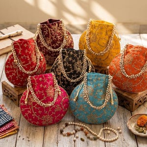 May include: A collection of six round drawstring pouches in red, maroon, black, yellow, orange, and turquoise. Each pouch features intricate gold floral embroidery and a decorative handle made of multiple strands of white faux pearls.