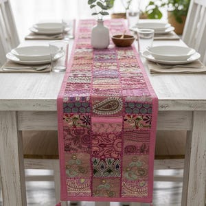 May include: A pink patchwork table runner with a paisley design and tassels. The runner is placed on a white wooden table set for a meal, with white plates, glasses, and silverware. A white vase with greenery is in the centre.