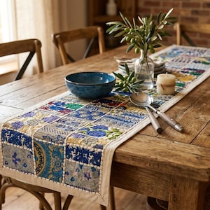 May include: A rectangular table runner featuring a patchwork design of embroidered fabric squares in shades of blue, green, and gold. The runner has a cream-coloured border and is decorated with intricate floral and geometric patterns throughout.