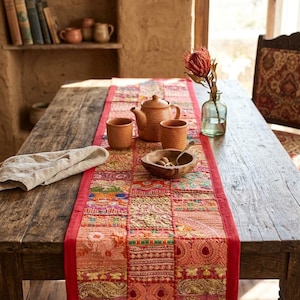 May include: A rectangular table runner featuring a patchwork design of various embroidered fabric squares in shades of red, orange, pink, and gold. The runner has a solid red border and is placed on a wooden table with a teapot and two cups.