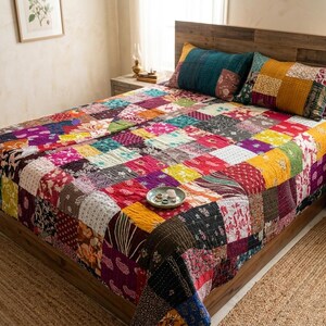 Puede incluir: Una colcha de patchwork que presenta una variedad de cuadrados de tela coloridos con diversos patrones florales, de cachemira y geom&eacute;tricos en tonos rojos, amarillos, morados, azules y verdes, cubriendo una cama con dos almohadas rectangulares a juego.