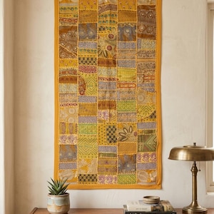 Puede incluir: Un tapiz rectangular con un diseño de patchwork compuesto por varios cuadrados de tela en tonos amarillo, dorado, verde y marrón. El tejido presenta bordados intrincados, pequeños detalles de espejo y diversos patrones geométricos y florales.