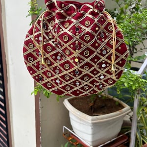 Könnte beinhalten: Eine bordeauxfarbene Samt-Beuteltasche mit goldener Stickerei und Spiegelakzenten. Die Tasche hat einen Kordelzugverschluss und Perlenhenkel. Das Design umfasst ein geometrisches Muster mit kreisförmigen und rautenförmigen Formen. Die Tasche hängt.