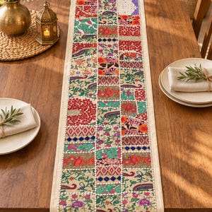 May include: A rectangular table runner featuring a patchwork design of embroidered fabric squares in vibrant colors including red, green, pink, and gold. The runner has a cream-colored border and is decorated with floral patterns and small mirrors.