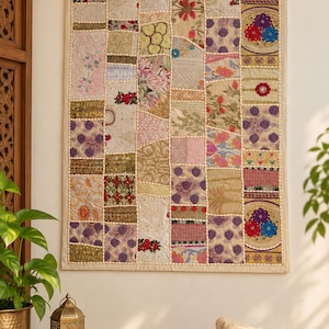 Op de afbeelding: Een rechthoekige wandhanger met een patchworkontwerp van diverse geborduurde stoffen vierkanten in tinten crème, goud, paars, rood en groen. Het textiel is omrand met een lichtbeige bies en versierd met kleine witte kraaltjes.