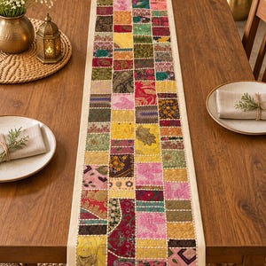 May include: A long rectangular table runner featuring a patchwork design of various fabric squares in vibrant colors including red, yellow, pink, green, and gold. The runner has a cream-colored border and visible white stitching between squares.