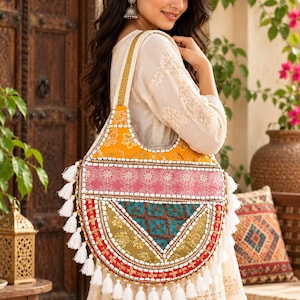 May include: A semi-circular shoulder bag featuring a patchwork design of yellow, pink, teal, and olive green fabric panels. The bag is decorated with white beaded trim, gold metallic accents, and a series of white tassels along the bottom edge.