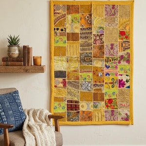 May include: A colorful patchwork wall hanging with a yellow border. The squares feature various patterns and textures in shades of gold, green, orange, and pink. It hangs on a cream-colored wall, adding a bohemian touch to the room.