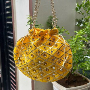 K&ouml;nnte beinhalten: Eine gelbe, runde Handtasche mit Kordelverschluss und Perlenhenkeln. Die Tasche ist mit goldener Stickerei, kleinen Spiegeln und goldenen Verzierungen verziert. Die Tasche h&auml;ngt vor einer gr&uuml;nen Pflanze.