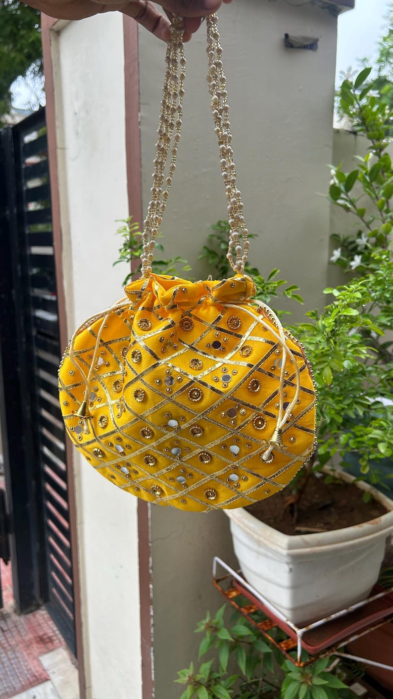 K&ouml;nnte beinhalten: Eine gelbe, runde Handtasche mit Kordelverschluss und Perlenhenkeln. Die Tasche ist mit goldener Stickerei, kleinen Spiegeln und goldenen Verzierungen verziert. Die Tasche h&auml;ngt vor einer gr&uuml;nen Pflanze.