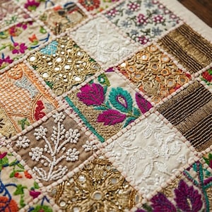 May include: A textile wall hanging featuring a patchwork design of square panels. Each square displays intricate embroidery, metallic gold thread work, small reflective mirrors, and white beadwork in floral and geometric patterns on fabric.