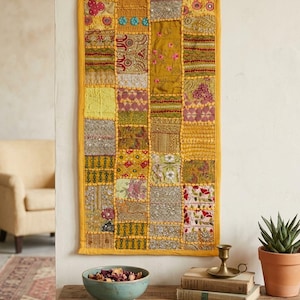 Op de afbeelding: Een rechthoekige wandhanger met een patchworkontwerp van diverse stoffen vierkanten in tinten geel, goud, roze en groen. Het textiel bevat ingewikkeld borduurwerk, kralenwerk en bloemmotieven, allemaal omrand door een frame van effen gele stof.