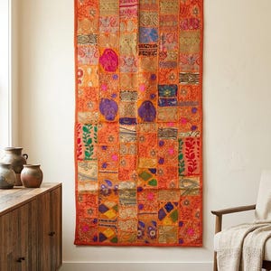 Op de afbeelding: Een rechthoekige wandhanger met een patchworkontwerp van diverse oranje stoffen vierkanten. Het stuk bevat uiteenlopende texturen, metallic gouden borduursels en kleurrijke bloem- en geometrische patronen die in een raster zijn gestikt.