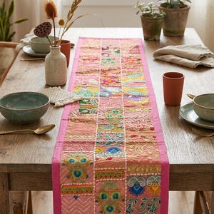Pode incluir: Um caminho de mesa retangular com um design de patchwork de quadrados de tecido rosa. Cada quadrado &eacute; decorado com bordados intrincados e multicoloridos, incluindo padr&otilde;es florais e geom&eacute;tricos, com uma borda rosa s&oacute;lida.