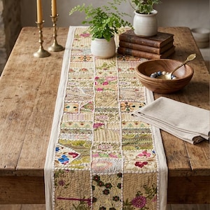 Pode incluir: Um caminho de mesa patchwork com padrões florais em tons de bege, rosa e verde, numa mesa de madeira rústica. A imagem inclui uma tigela de madeira, plantas e castiçais de latão. O caminho de mesa é feito de quadrados de tecido variados.