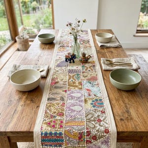 May include: A rectangular table runner featuring a patchwork design of embroidered fabric squares in various colors including gold, pink, blue, and purple. The runner is placed on a wooden table with four ceramic bowls and gold-toned cutlery.