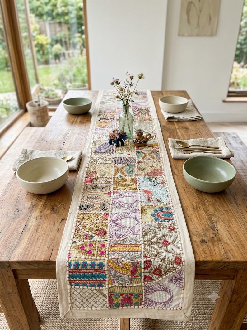 May include: A rectangular table runner featuring a patchwork design of embroidered fabric squares in various colours including gold, pink, blue, and purple. The runner is placed on a wooden table with four ceramic bowls and gold-toned cutlery.