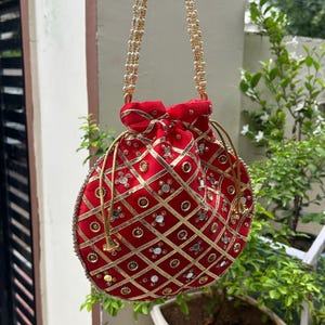 K&ouml;nnte beinhalten: Eine rote, runde Beuteltasche mit goldenen Verzierungen und einem goldperlenbesetzten Riemen. Die Tasche hat ein Steppmuster mit goldenen Zierleisten und kleinen, reflektierenden Akzenten. Die Tasche h&auml;ngt vor einem gr&uuml;nen, bl&auml;ttrigen Hintergrund.